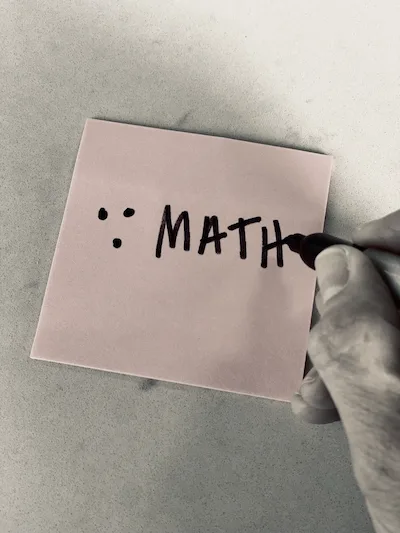 A pink notepad with a hand holding a pen that just finished wirting the math ematical symbol for the word 'therefore' and the word MATH in capitals.