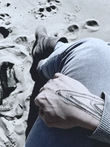 A hand with a graphical tattoo on it, resting on a lap of a person whose legs are in the sand.