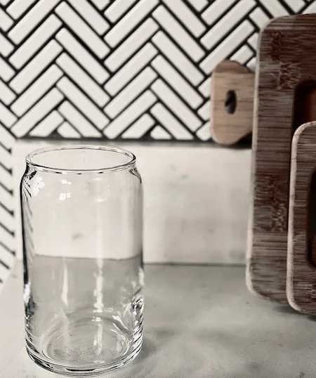 An empty glass on a counter with herringbone pattern on a wall in the background and the edges of a few wooden cutting boards