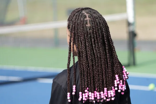 The view of the back of a person's head who has long braids with four pink beads at the end of each braid. They are wearing a black jacket and the ground is blue.