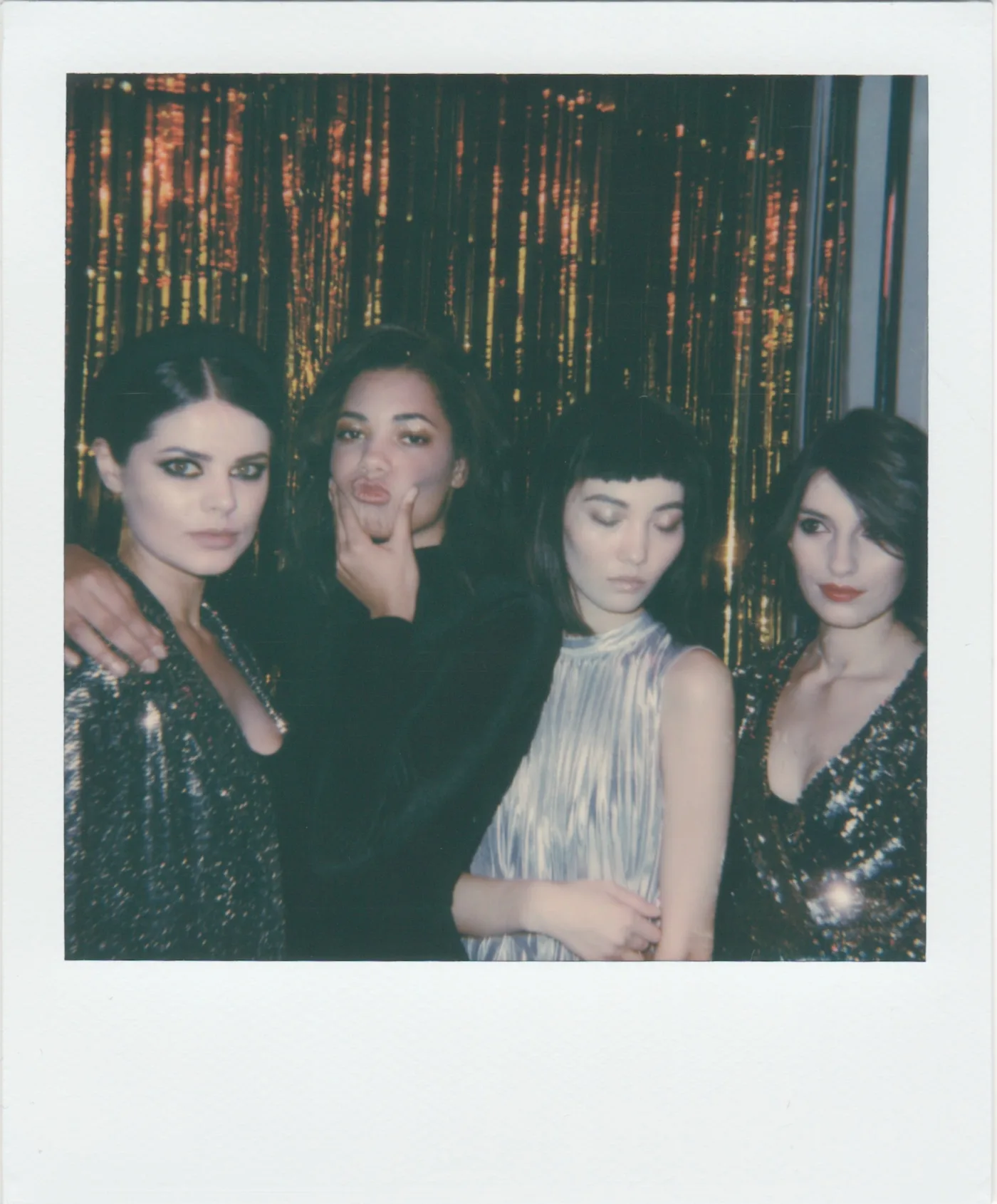 four brunettes in cocktail dresses pose in front of reddish-gold glittery curtain in a poloroid picture