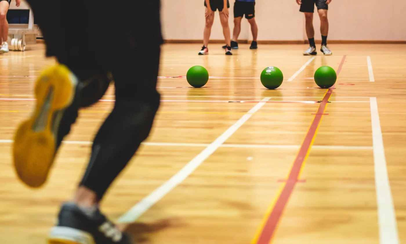 three green balls in the middle of the frame on olines as they roll on a gym floor. Bottom of torso and legs in foreground and three more people in far background.