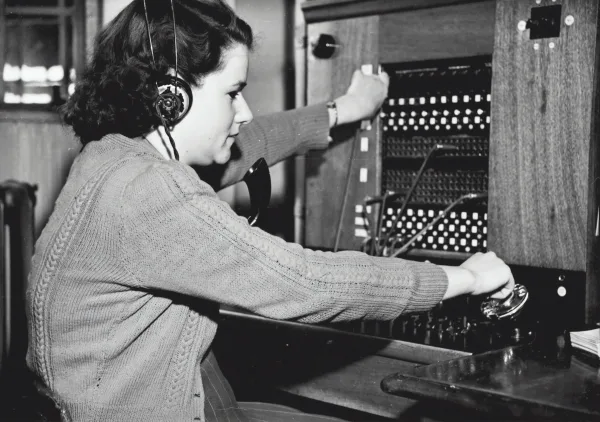a vintage black and white photo of a woman with a headset on who is turning knobs on a phone operator station.