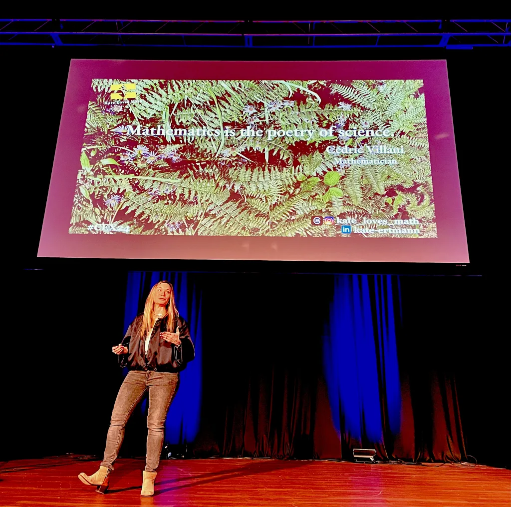 Mathemtician Kate Ertmann in foreground on left side, speaking from a stage with a projected image above that shows a quote, placed over a background of green fern leaves, from mathematician Cedric Villani that eads: Mathematics is the poetry of science.